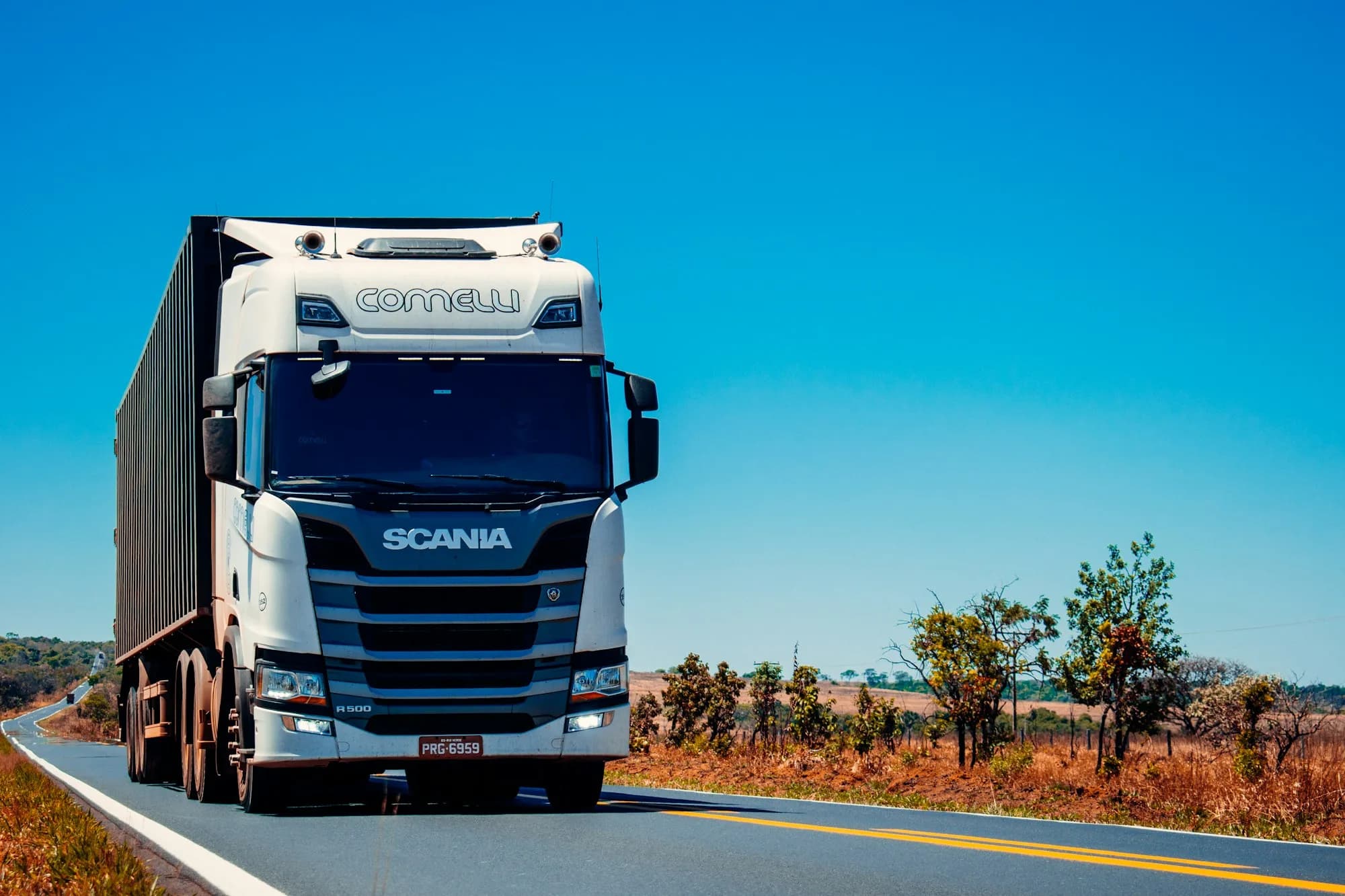 Biała ciężarówka Scania z naczepą jedzie drogą krajową w słoneczny dzień.